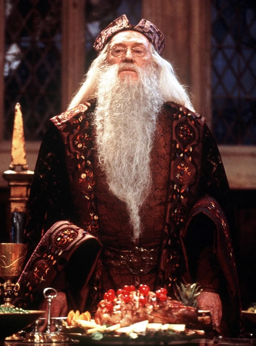 Richard Harris as Dumbledore presiding over a Hogwarts feast in an embroidered burgundy-and-gold brocade robe and matching hat, a glazed ham studded with cherries in front of him
